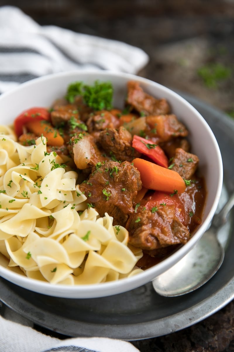 Close-up of a ladle lifting tender Hungarian Goulash beef chunks dripping with glossy paprika-red sauce over a bowl of buttered egg noodles, with a sour cream swirl and fresh dill garnish