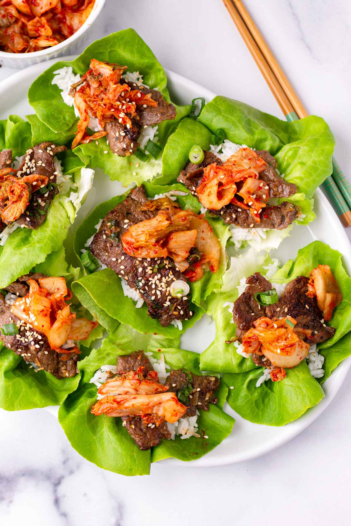 Korean BBQ lettuce wrap ingredients flat lay with marinated beef, butter lettuce cups, colorful pickled vegetables, gochujang sauce, and sesame seeds on a white marble surface
