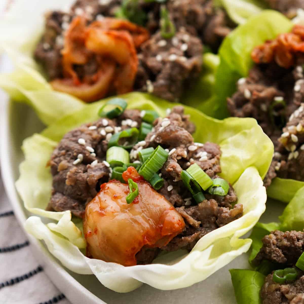 Close-up of a Korean BBQ lettuce wrap being held, showing caramelized beef, pickled vegetables, sesame seeds, and a drizzle of gochujang sauce spilling over the edge of a crisp butter lettuce cup