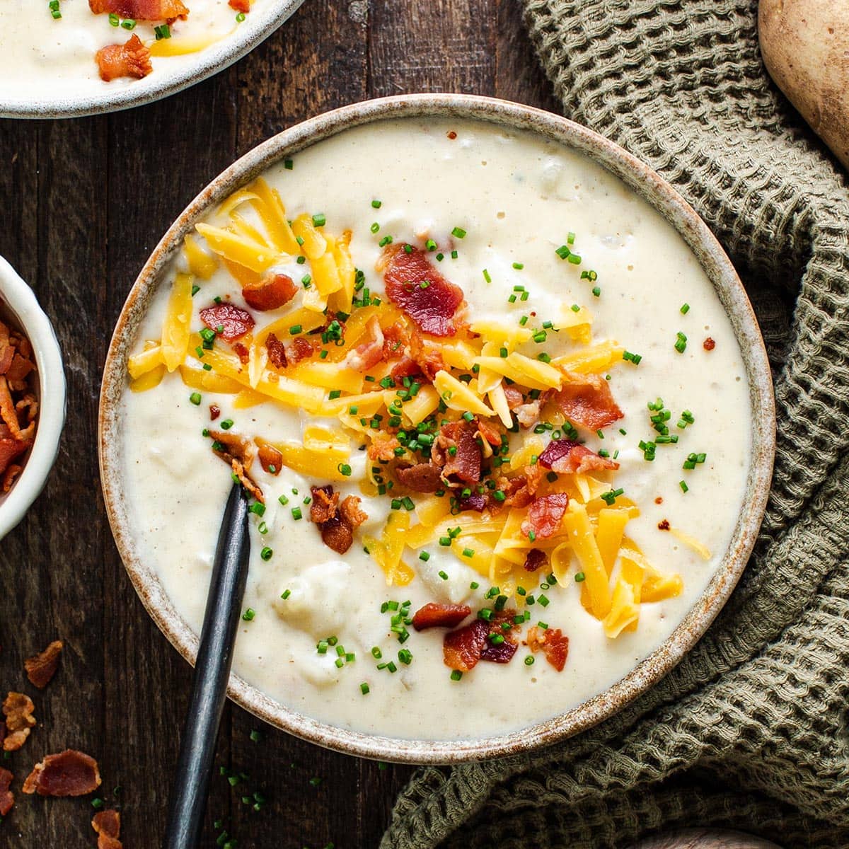 Overhead flat-lay of loaded baked potato soup ingredients: diced russet potatoes, strips of thick-cut bacon, shredded sharp cheddar, sour cream, butter, chicken broth, and fresh chives arranged on a marble surface