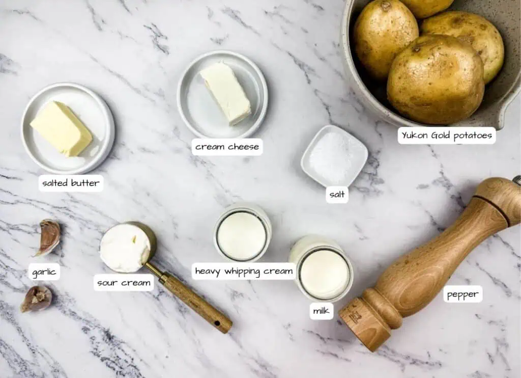 Overhead flat-lay of make ahead mashed potato ingredients including russet potatoes, cream cheese, butter, heavy cream, garlic, and chives arranged on a marble surface