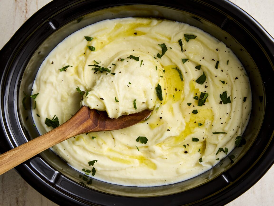 Close-up of creamy make ahead mashed potatoes in a slow cooker with melted butter pooled in the center, garnished with fresh chives and a pinch of flaky salt