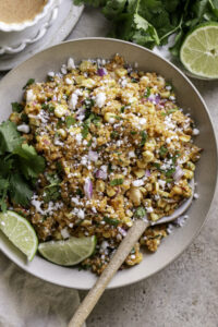 Mexican street corn quinoa salad in a wide bowl garnished with cotija cheese, fresh cilantro, chili powder, and lime wedges on a rustic wooden table