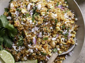 Mexican street corn quinoa salad in a wide bowl garnished with cotija cheese, fresh cilantro, chili powder, and lime wedges on a rustic wooden table