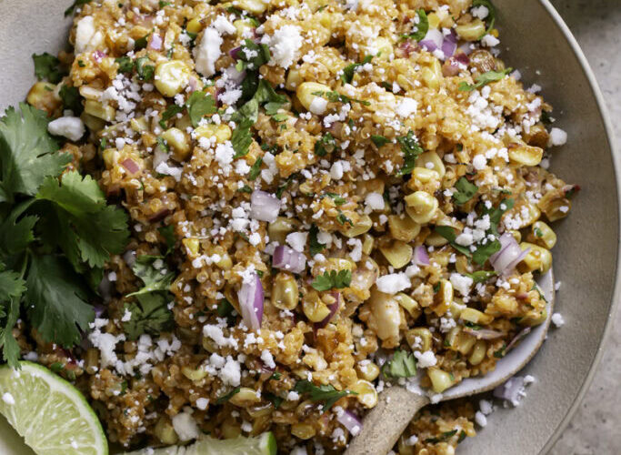 Mexican street corn quinoa salad in a wide bowl garnished with cotija cheese, fresh cilantro, chili powder, and lime wedges on a rustic wooden table