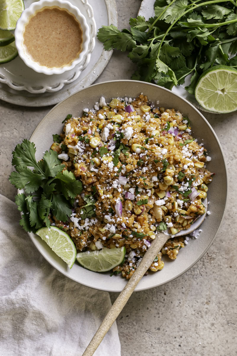 Close-up of Mexican street corn quinoa salad served in a terracotta bowl with charred corn kernels visible, cotija cheese crumbles, fresh cilantro, and a lime wedge on the side