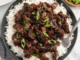 Mongolian Beef served over steamed white rice in a dark bowl, garnished with sliced green onions and sesame seeds, glossy brown sauce coating tender strips of flank steak