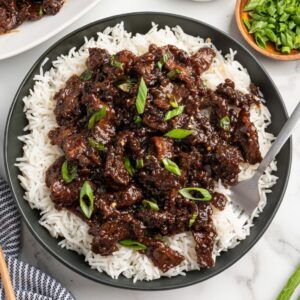 Mongolian Beef served over steamed white rice in a dark bowl, garnished with sliced green onions and sesame seeds, glossy brown sauce coating tender strips of flank steak