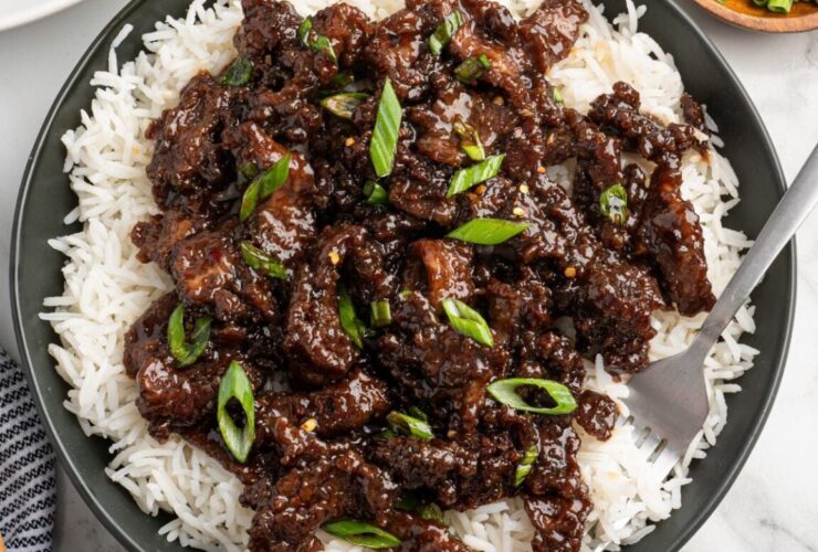 Mongolian Beef served over steamed white rice in a dark bowl, garnished with sliced green onions and sesame seeds, glossy brown sauce coating tender strips of flank steak