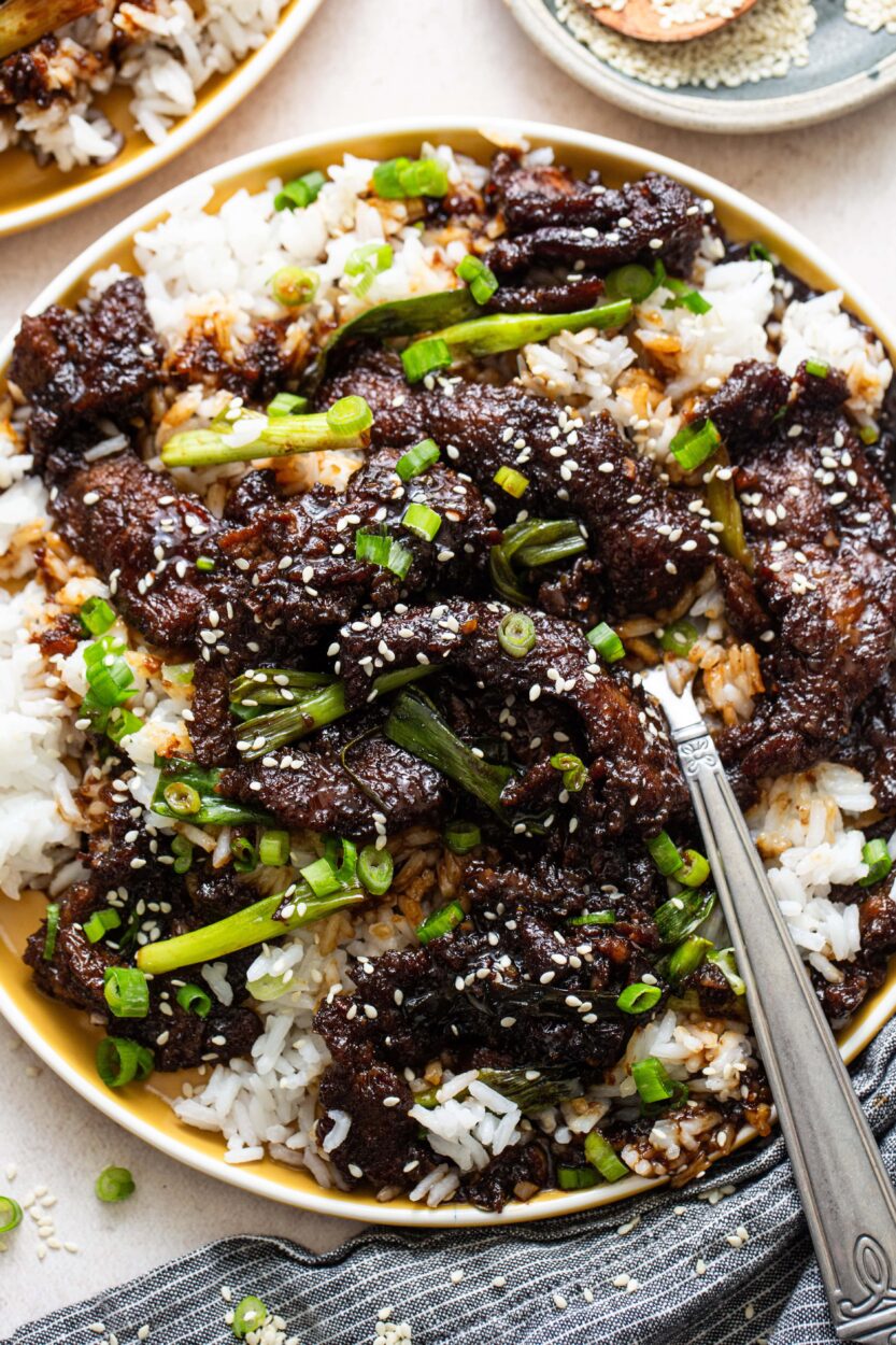 Overhead flat lay of Mongolian Beef ingredients including sliced raw flank steak, bowls of soy sauce, brown sugar, minced garlic and ginger, cornstarch, and green onions on a dark surface