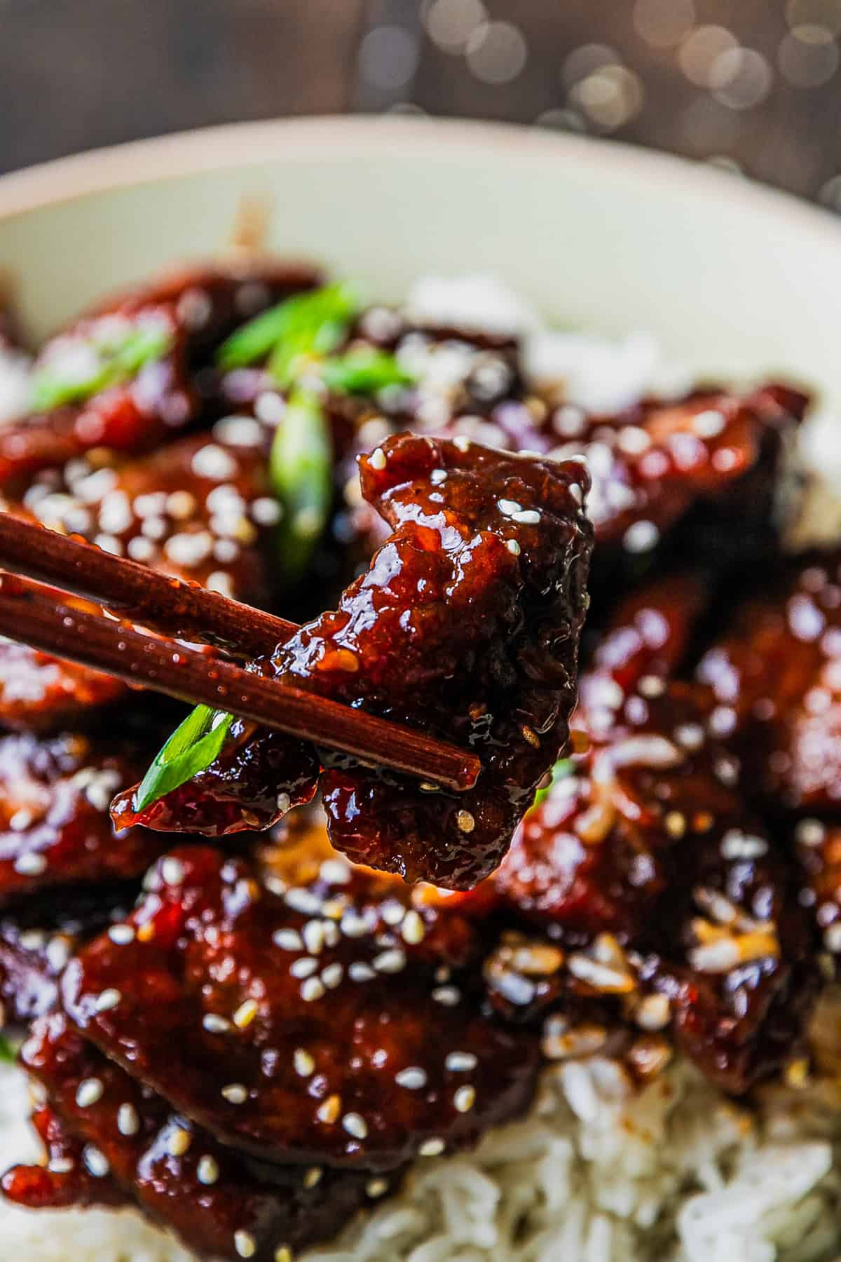 Close-up of Mongolian Beef in a serving bowl showing glossy dark brown sauce coating tender crispy-edged beef strips, green onion slivers and sesame seeds on top, steamed rice visible in background