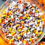 Monster Munch Halloween puppy chow in a large bowl with candy eyes, orange white chocolate drizzle, pretzels, and Halloween sprinkles on a dark spooky tablescape