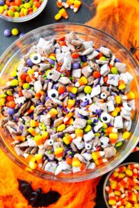Monster Munch Halloween puppy chow in a large bowl with candy eyes, orange white chocolate drizzle, pretzels, and Halloween sprinkles on a dark spooky tablescape
