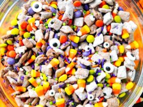 Monster Munch Halloween puppy chow in a large bowl with candy eyes, orange white chocolate drizzle, pretzels, and Halloween sprinkles on a dark spooky tablescape