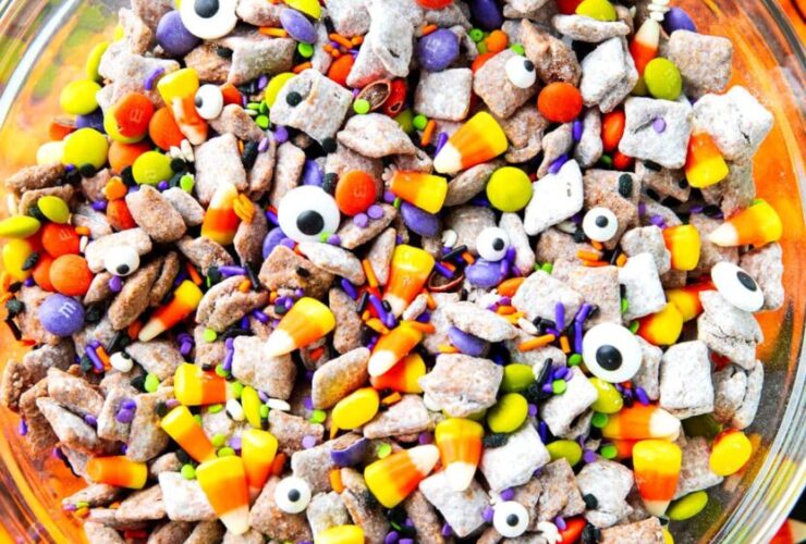 Monster Munch Halloween puppy chow in a large bowl with candy eyes, orange white chocolate drizzle, pretzels, and Halloween sprinkles on a dark spooky tablescape
