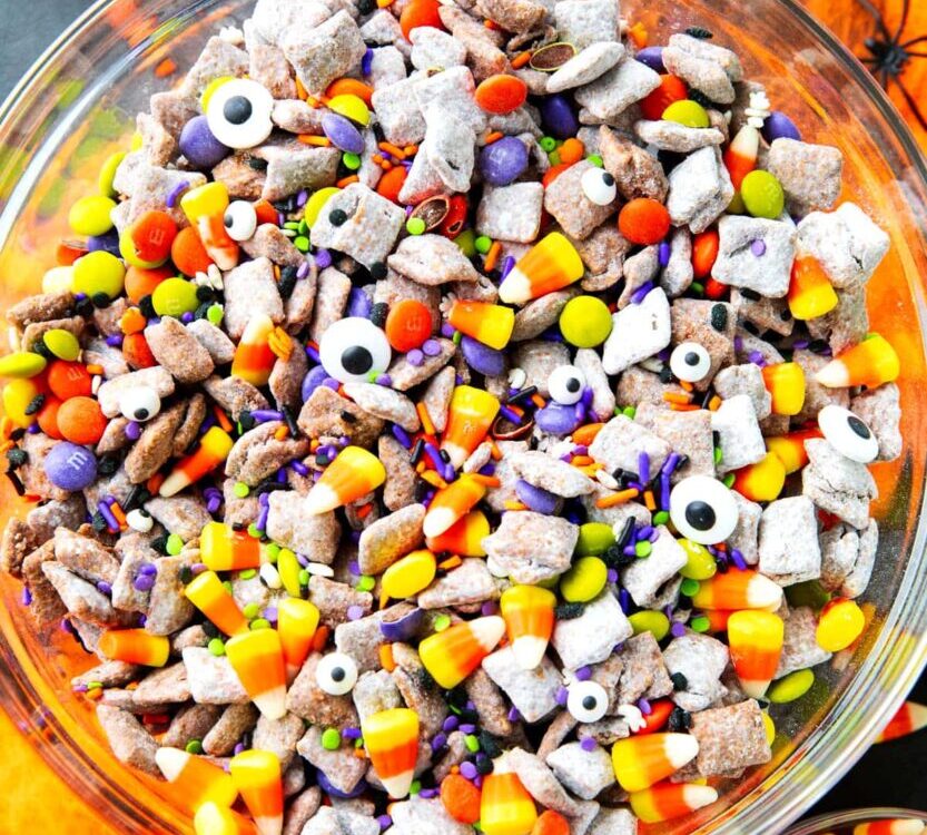 Monster Munch Halloween puppy chow in a large bowl with candy eyes, orange white chocolate drizzle, pretzels, and Halloween sprinkles on a dark spooky tablescape