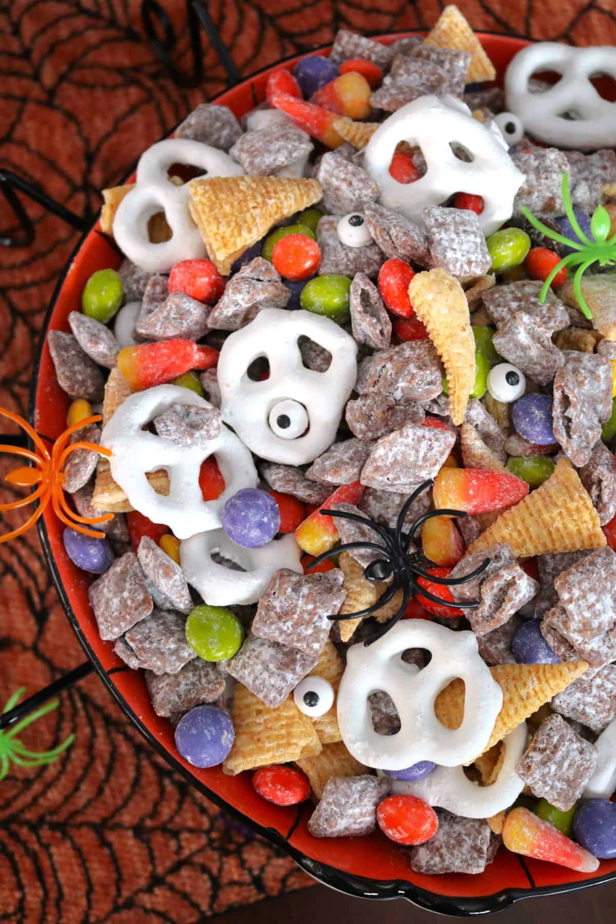 Flat lay of Monster Munch Halloween puppy chow ingredients including rice Chex cereal, pretzels, chocolate chips, white chocolate chips, peanut butter, candy eyes, orange food coloring, and Halloween sprinkles on a dark surface