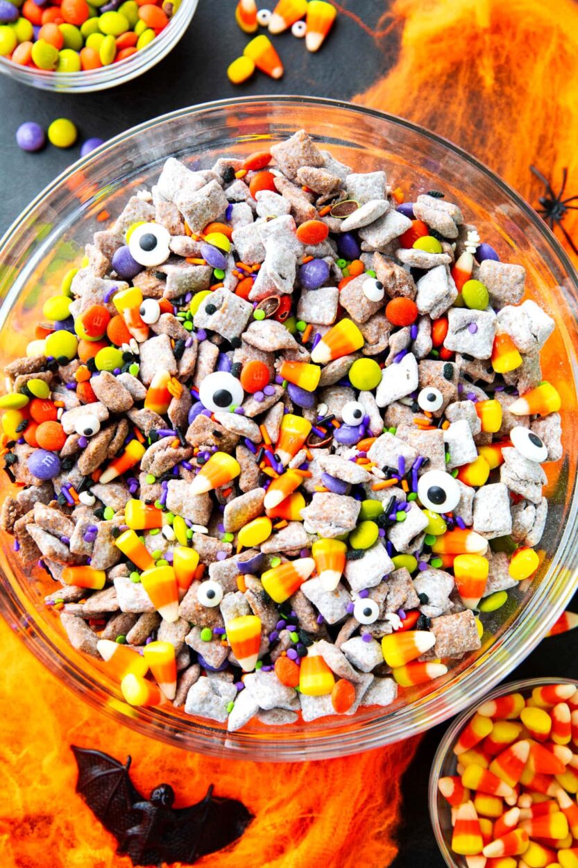 Close-up of Monster Munch Halloween puppy chow in a black serving bowl with candy eyes staring up, orange white chocolate drizzle visible, and Halloween sprinkles scattered throughout, moody Halloween lighting
