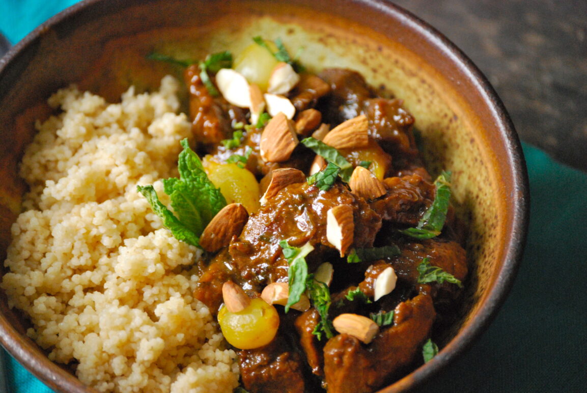 Moroccan lamb tagine served in a traditional clay tagine pot with couscous, garnished with fresh cilantro, slivered almonds, and golden raisins on a rustic wooden table