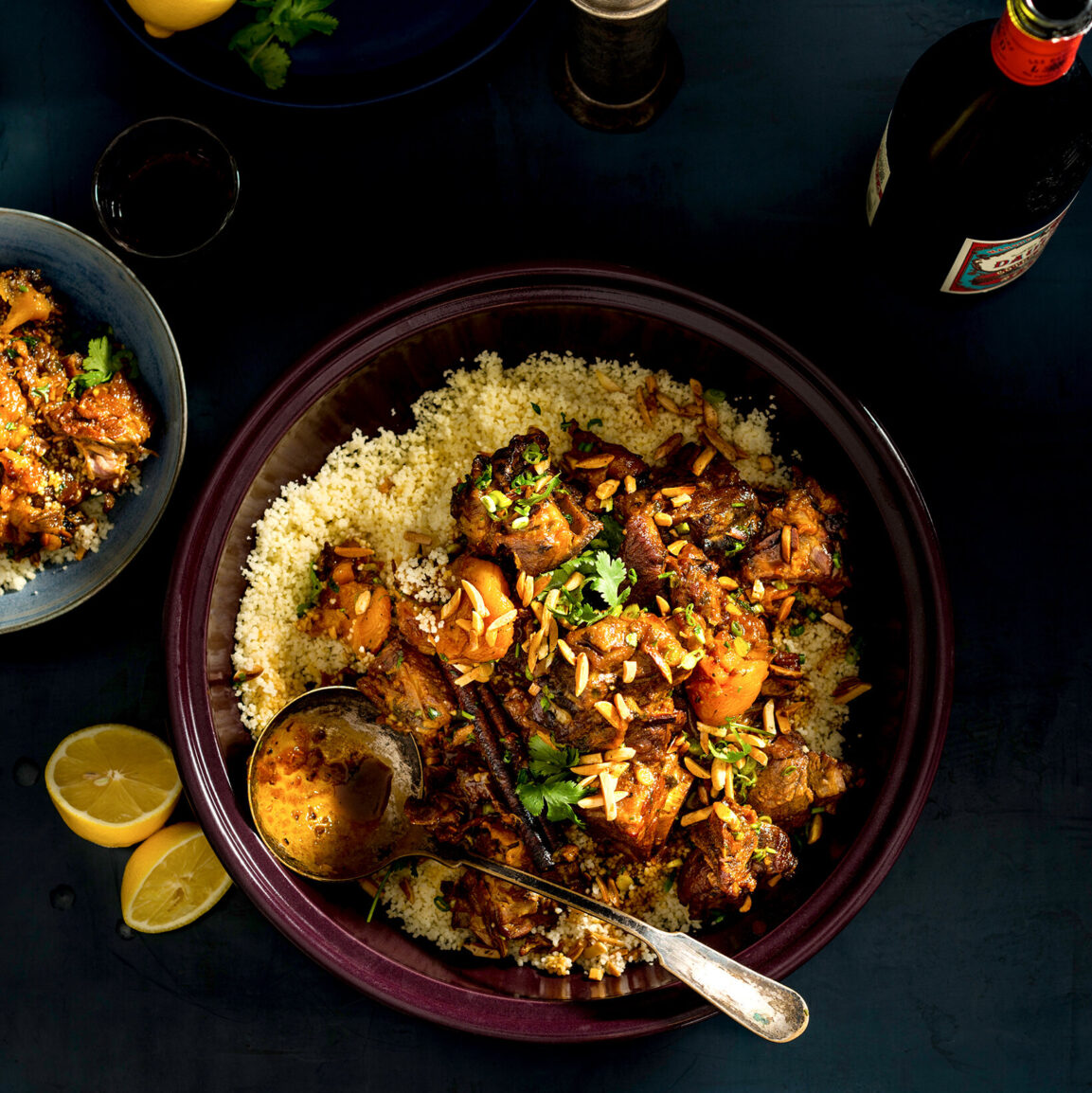 Overhead flat lay of Moroccan lamb tagine ingredients including lamb shoulder pieces, dried apricots, preserved lemon, whole spices, onions, tomatoes, and fresh herbs arranged on a dark stone surface