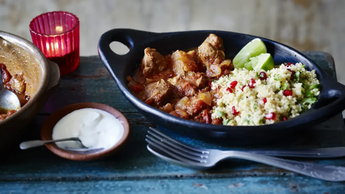 Close-up of Moroccan lamb tagine sauce-coated lamb pieces plated over fluffy couscous, topped with pomegranate seeds, fresh mint leaves, and a wedge of preserved lemon on a white ceramic plate