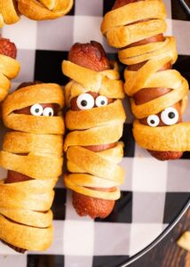 Platter of golden mummy hot dogs with mustard eyes on a dark Halloween-themed serving board, steam rising, beautifully styled