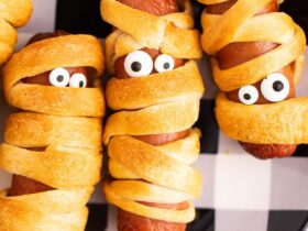 Platter of golden mummy hot dogs with mustard eyes on a dark Halloween-themed serving board, steam rising, beautifully styled