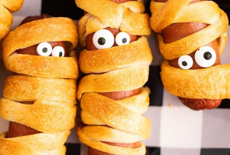 Platter of golden mummy hot dogs with mustard eyes on a dark Halloween-themed serving board, steam rising, beautifully styled
