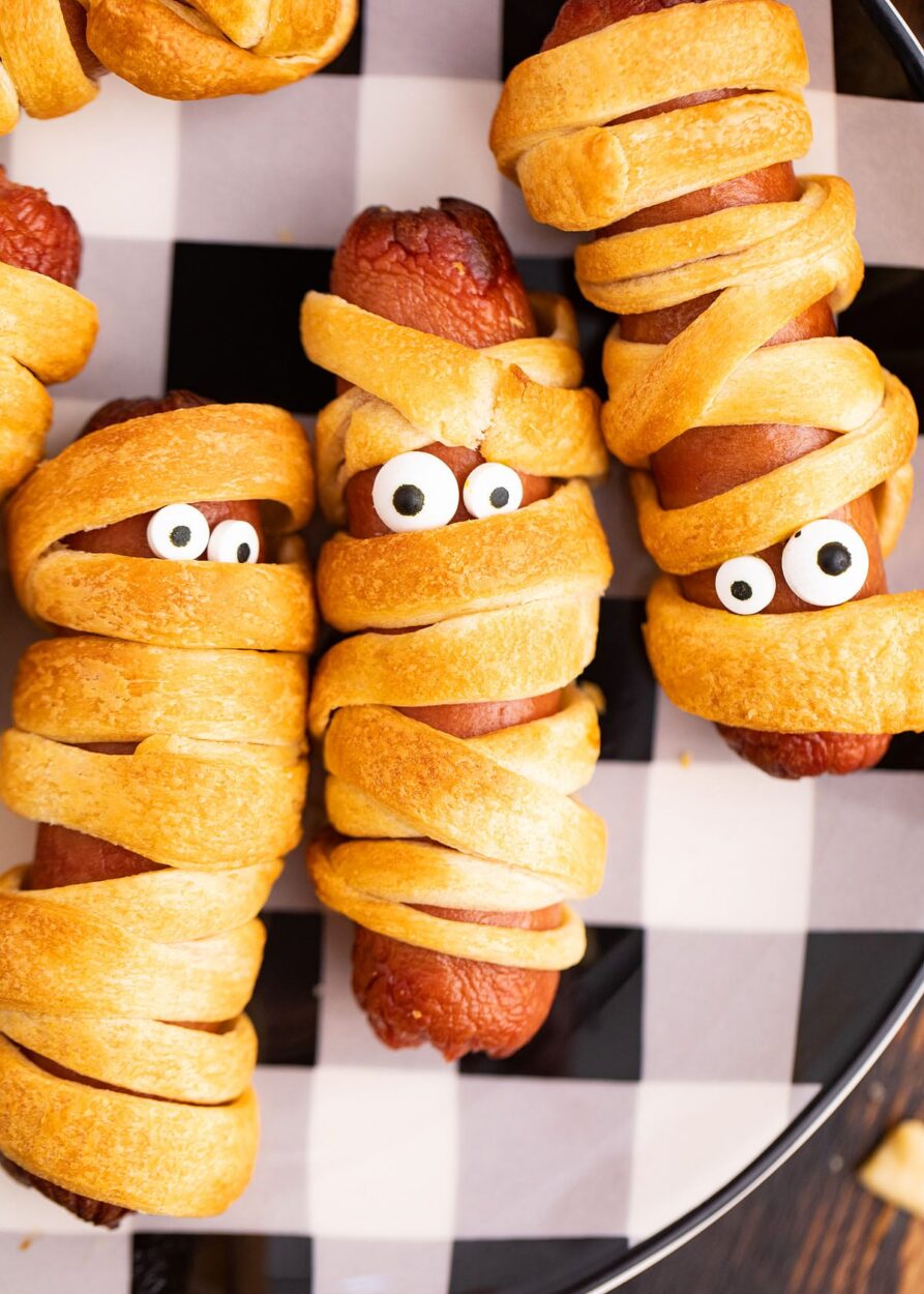 Platter of golden mummy hot dogs with mustard eyes on a dark Halloween-themed serving board, steam rising, beautifully styled