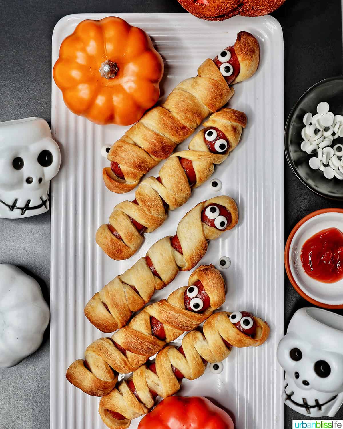 Overhead flat-lay of crescent dough strips being wrapped around hot dogs on a parchment-lined baking sheet, mustard bottle and googly candy eyes nearby