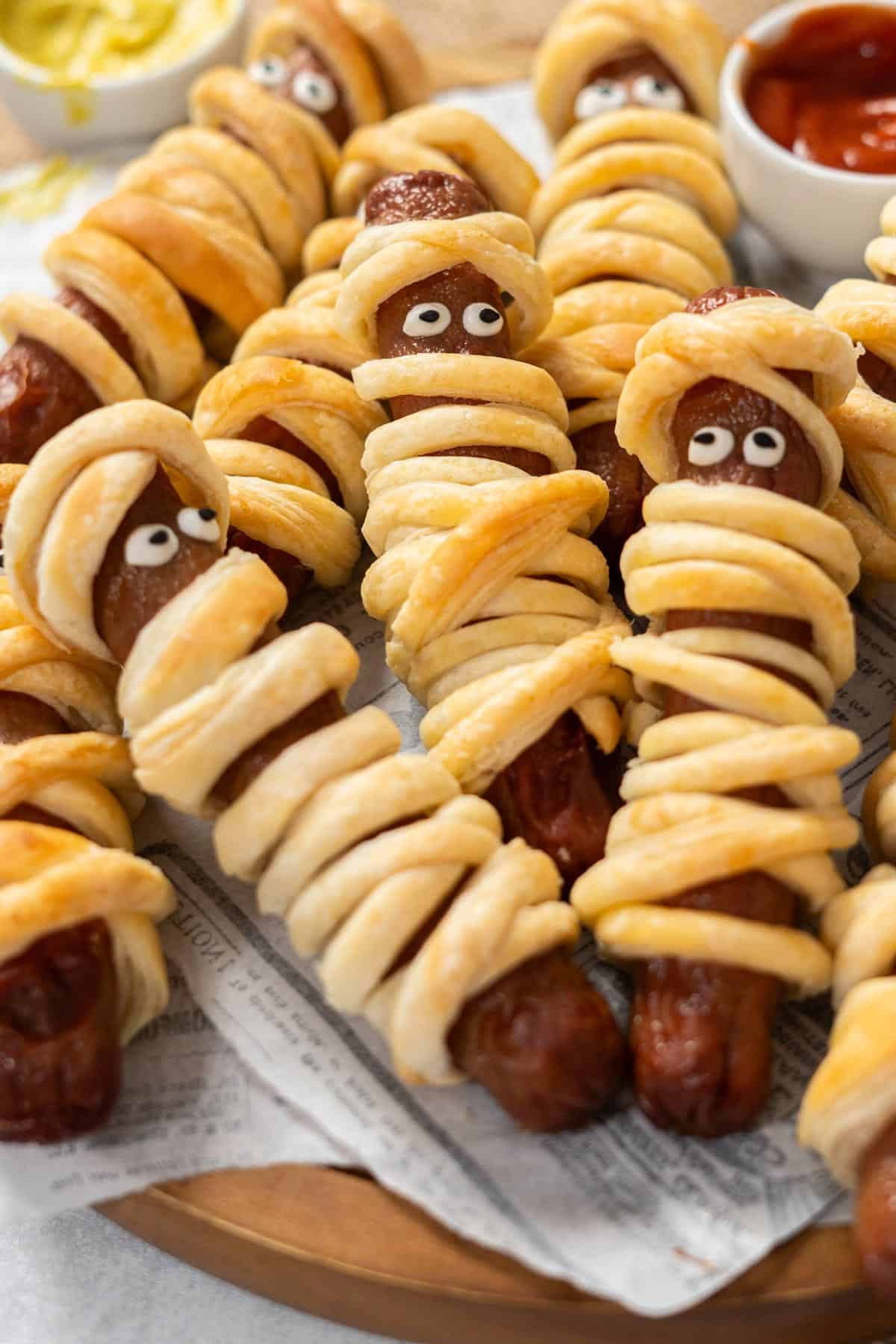Close-up of a single mummy hot dog with golden flaky crescent pastry and two mustard eyes, held up with a spooky Halloween backdrop, steam and shine visible