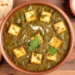 Palak Paneer served in a dark bowl with golden paneer cubes nestled in vibrant green spinach sauce, garnished with a swirl of cream and fresh ginger julienne, shot overhead on a wooden table with naan bread alongside
