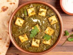 Palak Paneer served in a dark bowl with golden paneer cubes nestled in vibrant green spinach sauce, garnished with a swirl of cream and fresh ginger julienne, shot overhead on a wooden table with naan bread alongside