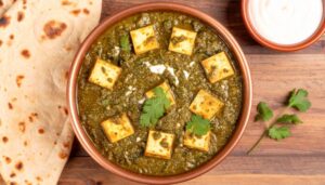 Palak Paneer served in a dark bowl with golden paneer cubes nestled in vibrant green spinach sauce, garnished with a swirl of cream and fresh ginger julienne, shot overhead on a wooden table with naan bread alongside
