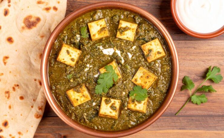 Palak Paneer served in a dark bowl with golden paneer cubes nestled in vibrant green spinach sauce, garnished with a swirl of cream and fresh ginger julienne, shot overhead on a wooden table with naan bread alongside