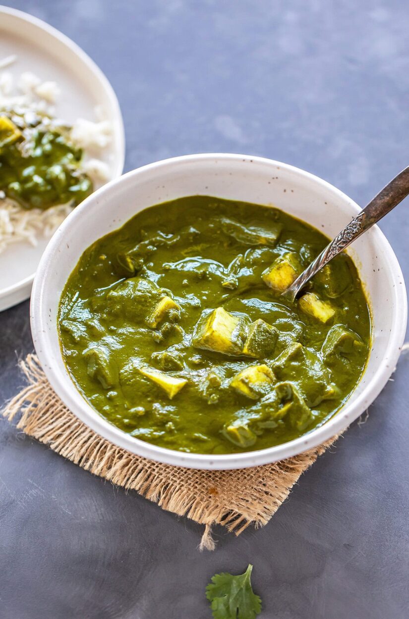 Overhead flat lay of Palak Paneer ingredients including fresh spinach, cubed paneer, whole spices in small bowls, fresh ginger, garlic, tomatoes, and a small bowl of cream arranged on a marble surface