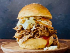 Pulled pork sandwiches piled high on toasted brioche buns with coleslaw and BBQ sauce dripping down the sides, shot on a wooden board with pickles and chips on the side