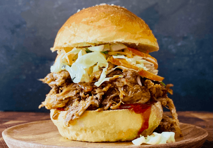 Pulled pork sandwiches piled high on toasted brioche buns with coleslaw and BBQ sauce dripping down the sides, shot on a wooden board with pickles and chips on the side