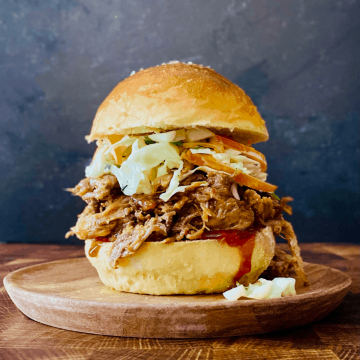 Pulled pork sandwiches piled high on toasted brioche buns with coleslaw and BBQ sauce dripping down the sides, shot on a wooden board with pickles and chips on the side
