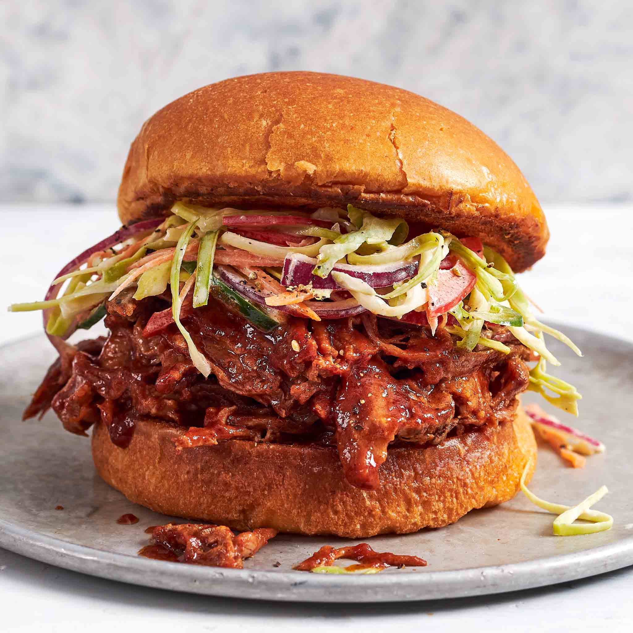 Overhead flat-lay of pulled pork ingredients including pork shoulder, spice rub in small bowls, BBQ sauce, apple cider vinegar, and brioche buns on a white surface