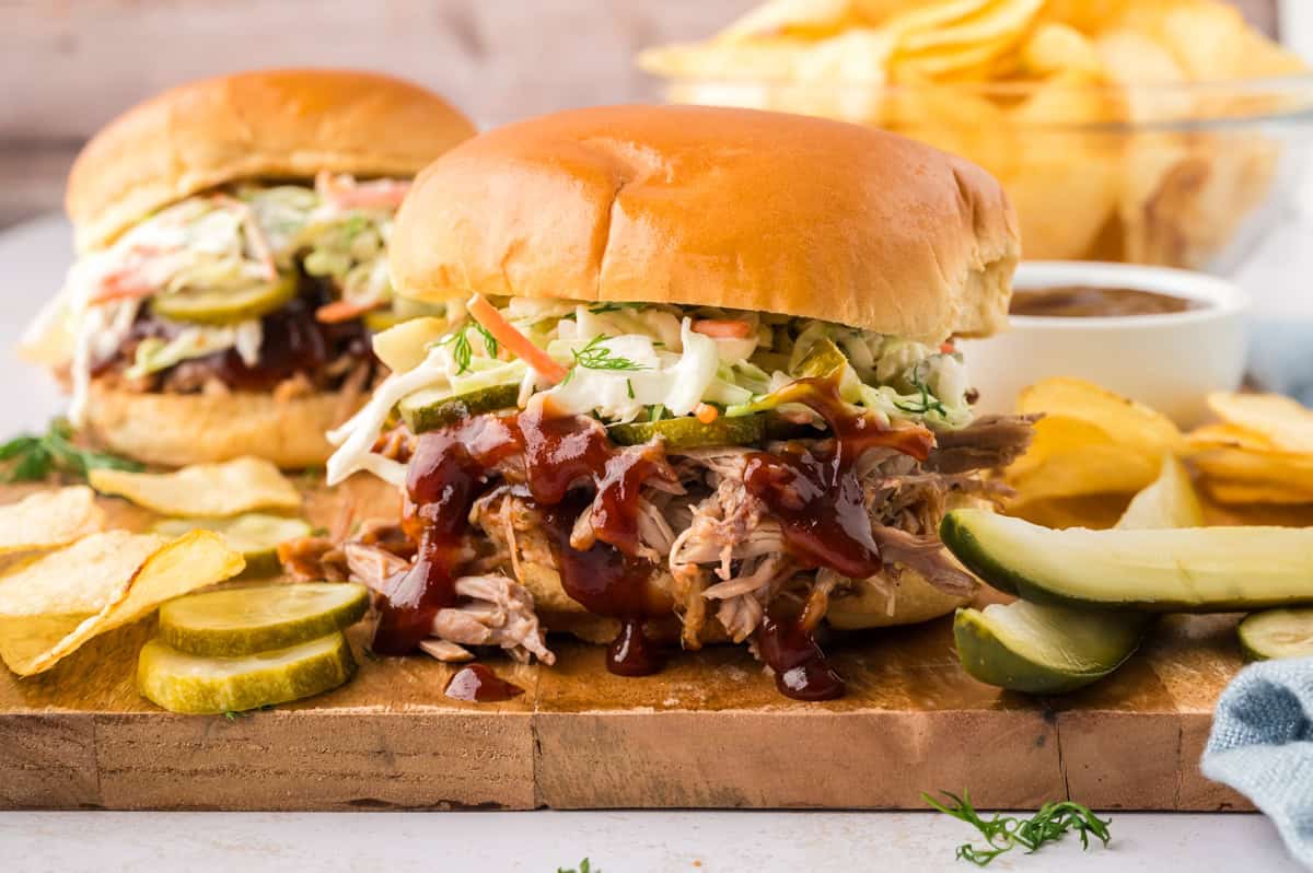 Close-up of a pulled pork sandwich being pulled apart to show the tender, saucy shredded pork and crispy coleslaw inside a toasted brioche bun, with BBQ sauce dripping onto the board
