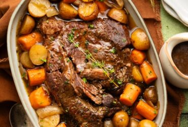 Slow cooker pot roast served on a large platter with tender carrots, potatoes, and onions, topped with glossy dark gravy, garnished with fresh thyme