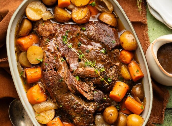 Slow cooker pot roast served on a large platter with tender carrots, potatoes, and onions, topped with glossy dark gravy, garnished with fresh thyme