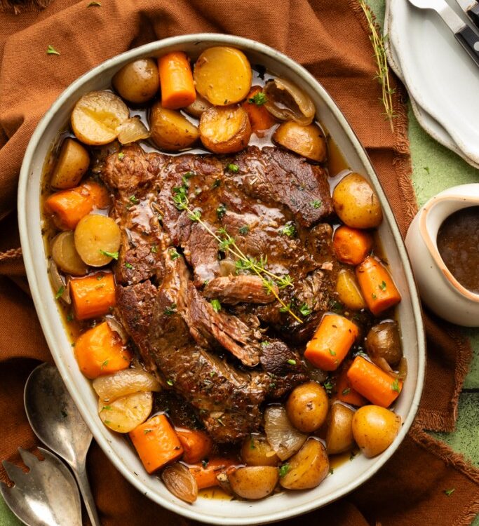 Slow cooker pot roast served on a large platter with tender carrots, potatoes, and onions, topped with glossy dark gravy, garnished with fresh thyme
