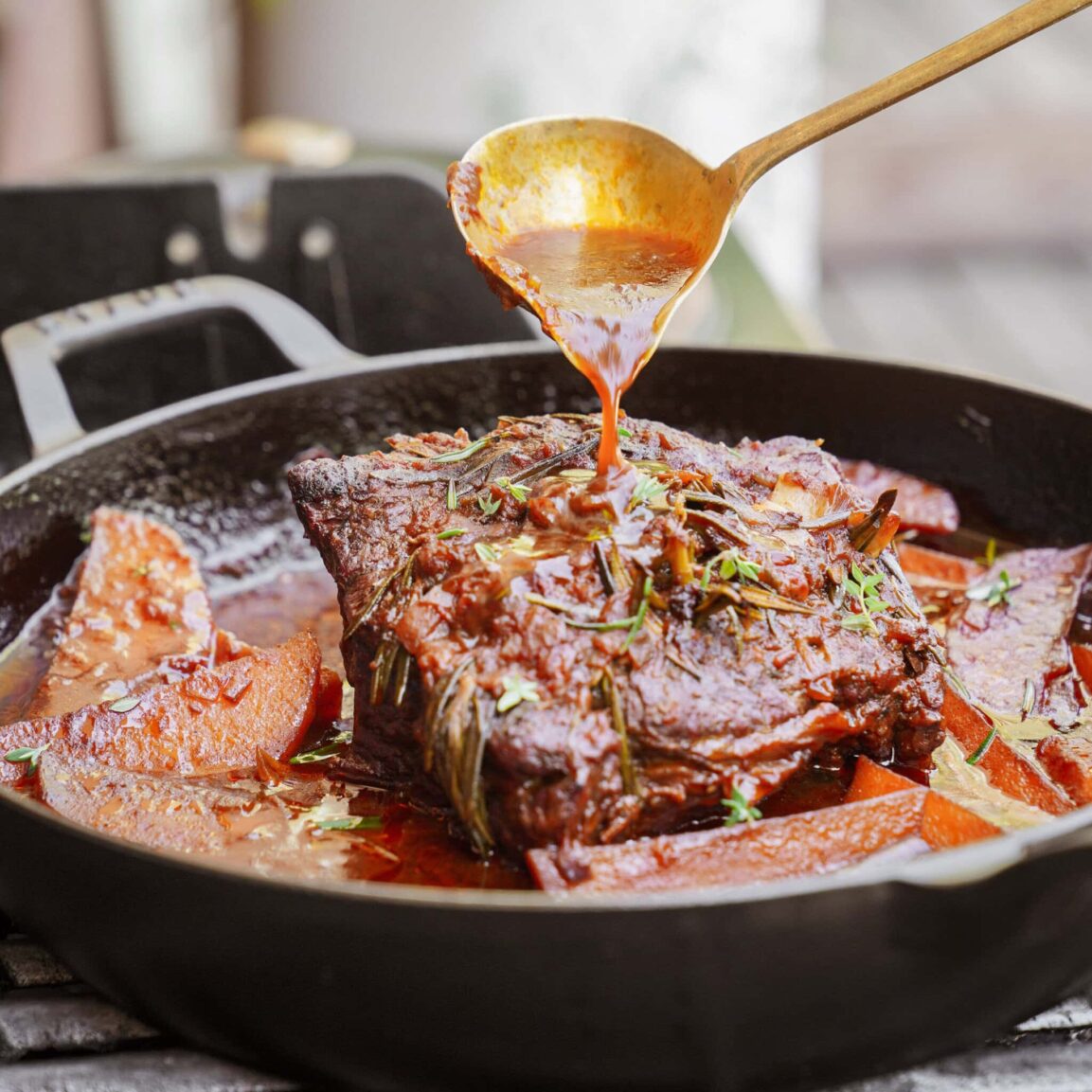 Chuck roast being seared in a cast iron skillet with a deep golden-brown crust, with bowls of chopped root vegetables, garlic, tomato paste, and fresh herbs arranged nearby