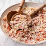 A large white serving bowl overflowing with creamy Southern coleslaw, garnished with a sprinkle of paprika and fresh parsley, shot on a rustic wooden picnic table with a checkered napkin alongside