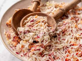 A large white serving bowl overflowing with creamy Southern coleslaw, garnished with a sprinkle of paprika and fresh parsley, shot on a rustic wooden picnic table with a checkered napkin alongside