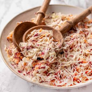 A large white serving bowl overflowing with creamy Southern coleslaw, garnished with a sprinkle of paprika and fresh parsley, shot on a rustic wooden picnic table with a checkered napkin alongside