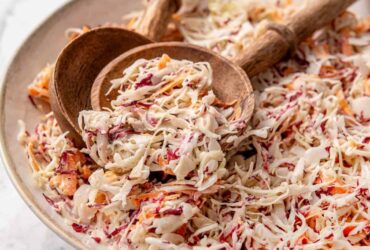 A large white serving bowl overflowing with creamy Southern coleslaw, garnished with a sprinkle of paprika and fresh parsley, shot on a rustic wooden picnic table with a checkered napkin alongside