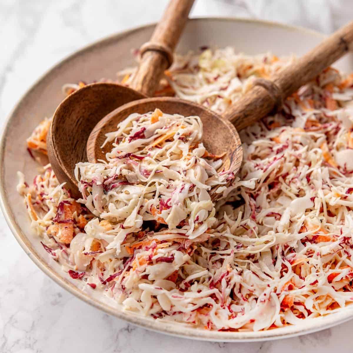 A large white serving bowl overflowing with creamy Southern coleslaw, garnished with a sprinkle of paprika and fresh parsley, shot on a rustic wooden picnic table with a checkered napkin alongside